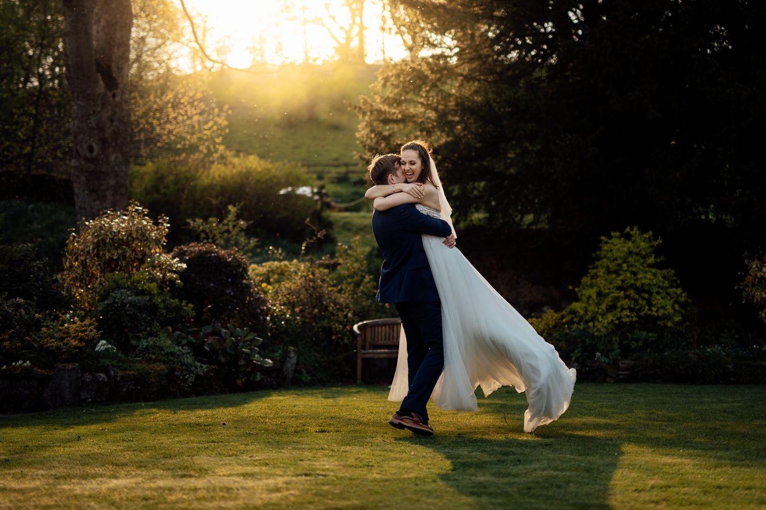 Couple walking through a scenic garden path at golden hour with dramatic sunlight filtering through trees.
