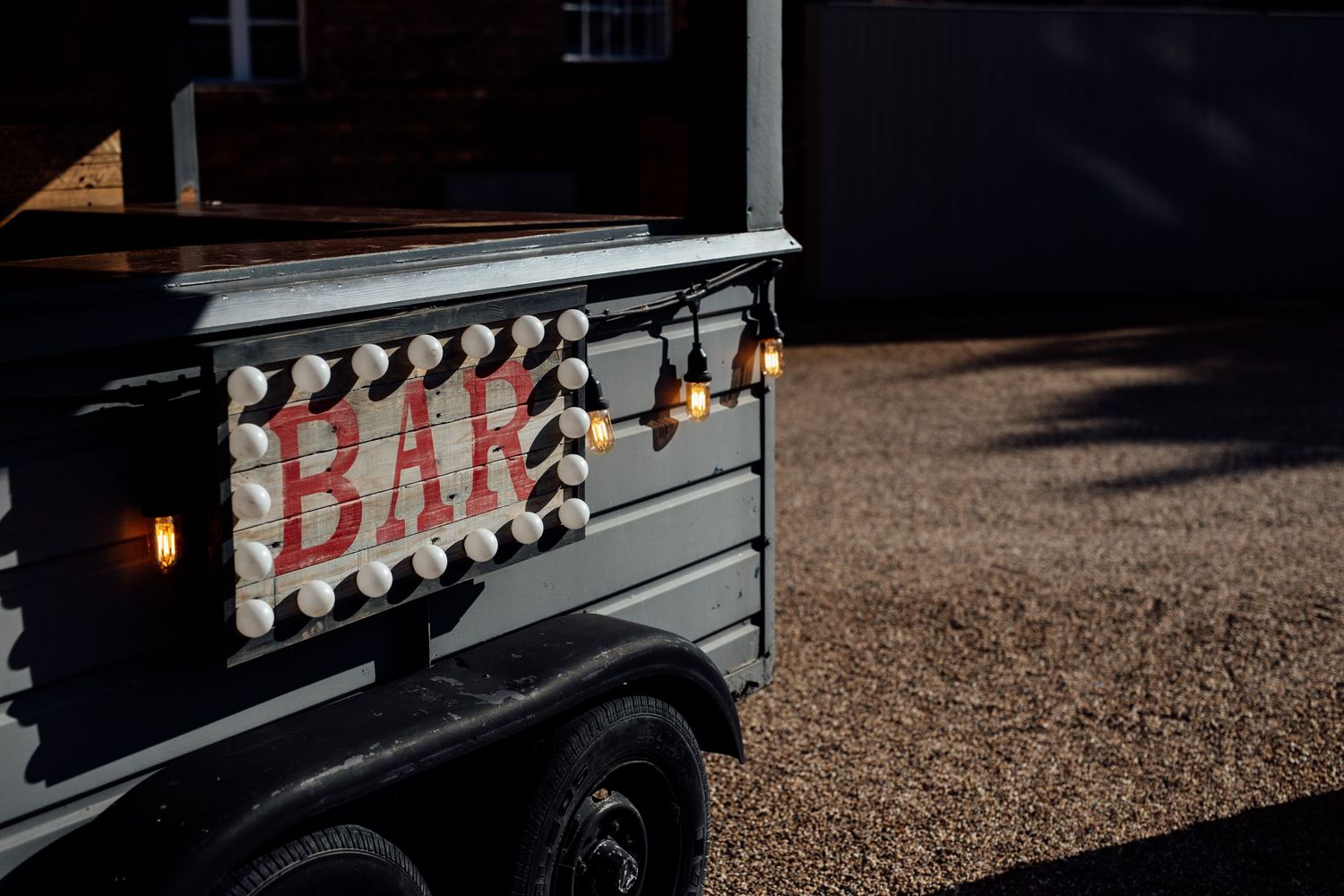 Black vintage trailer with illuminated 'BAR' sign at outdoor event.