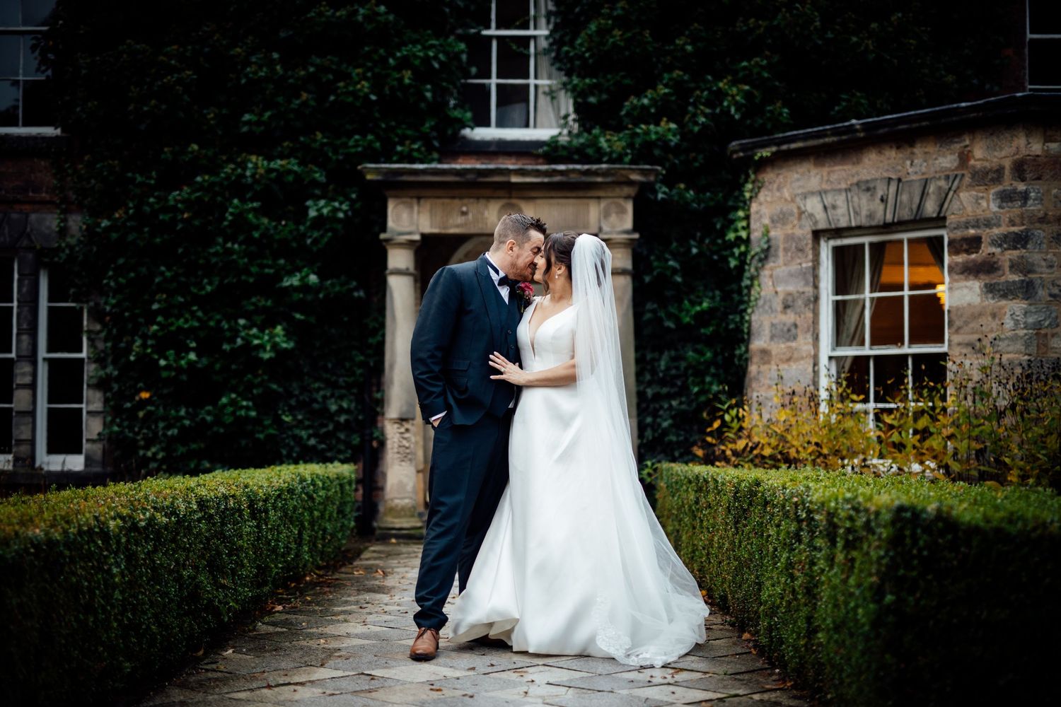 Romantic wedding portrait on stone pathway with historic building and manicured hedges.