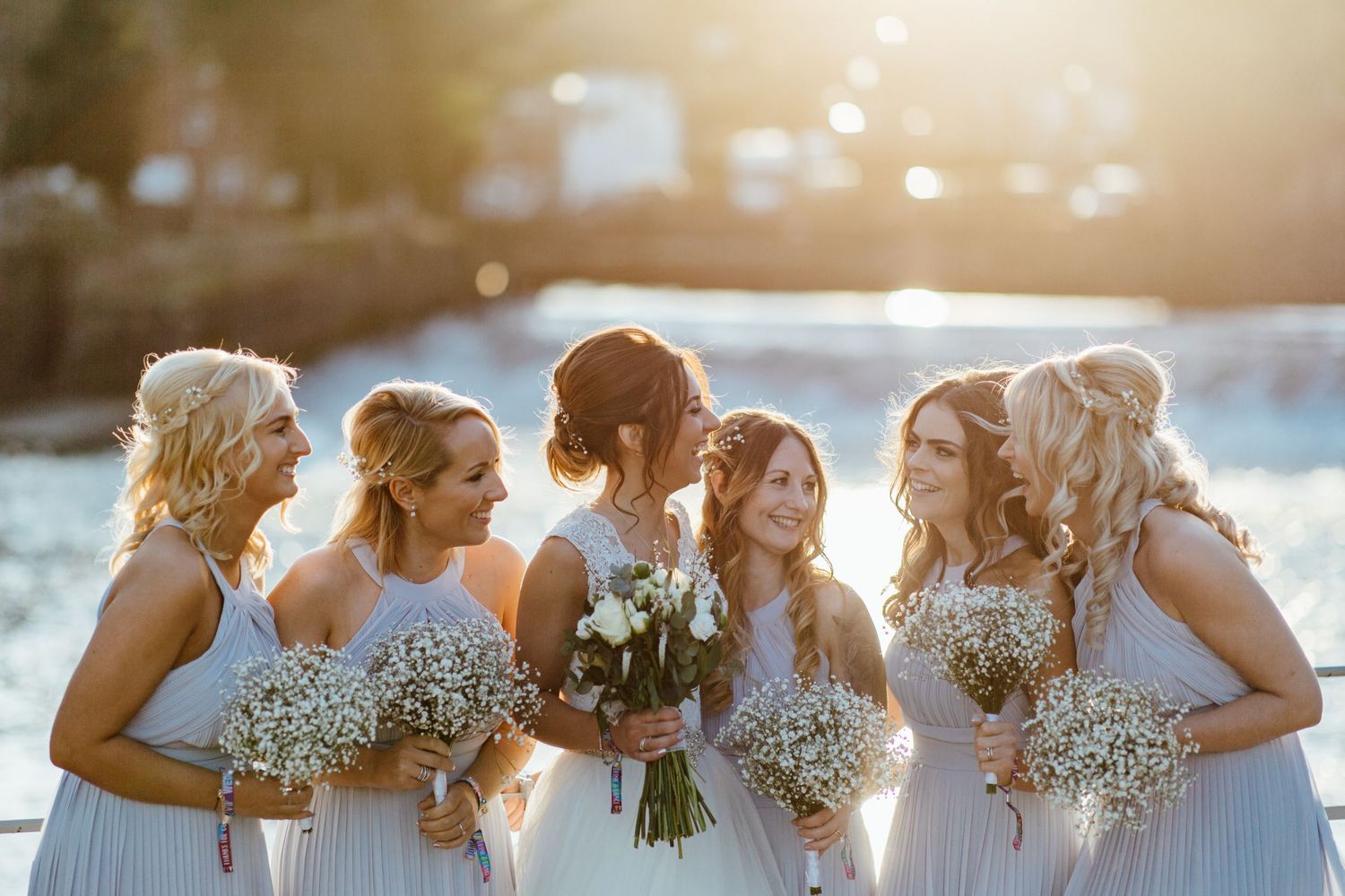 Bridesmaids in light colored dresses stand together holding bouquets at sunset.