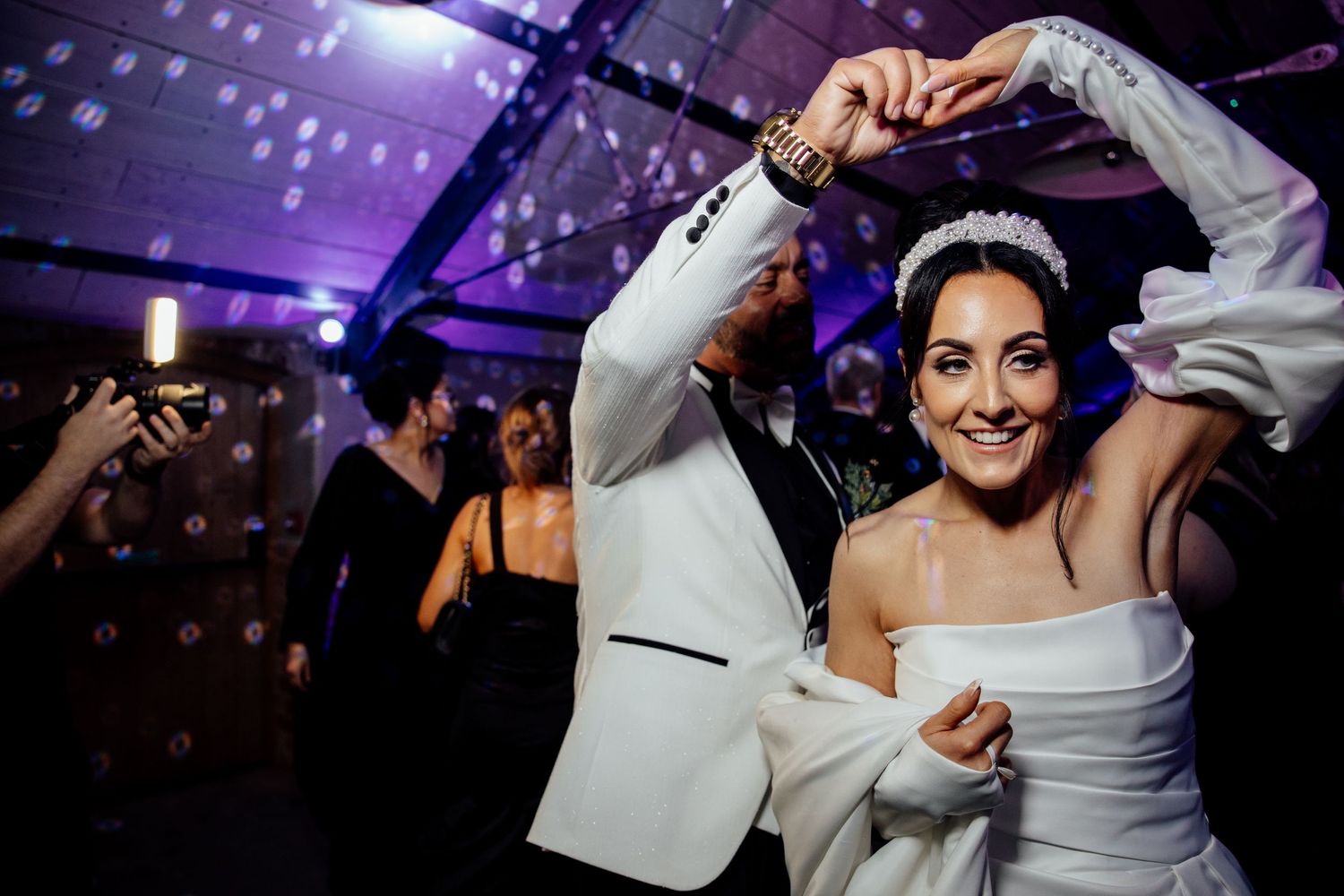 Wedding party dancing under purple lighting with festive atmosphere at reception.