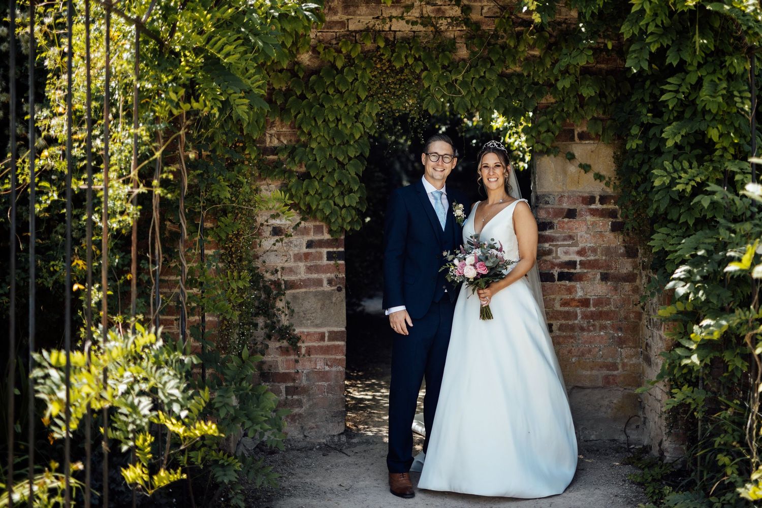 A couple poses under an ivy-covered brick archway in formal wedding attire.