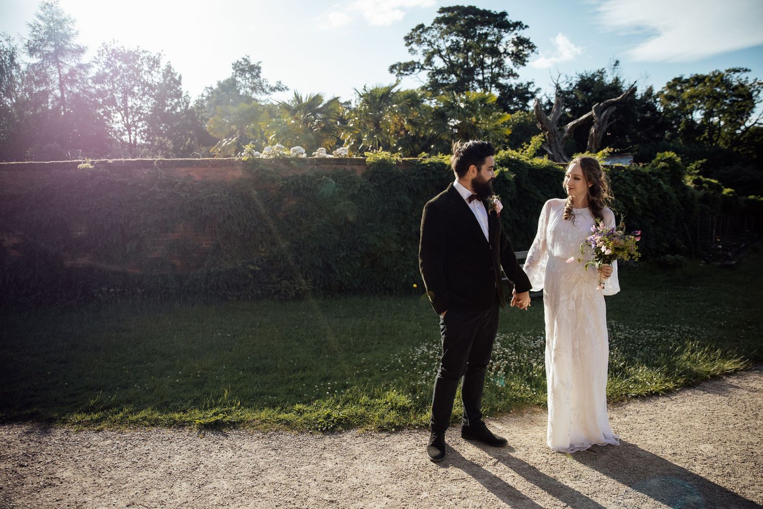 A couple in wedding attire walks together through a sunlit garden path during golden hour.