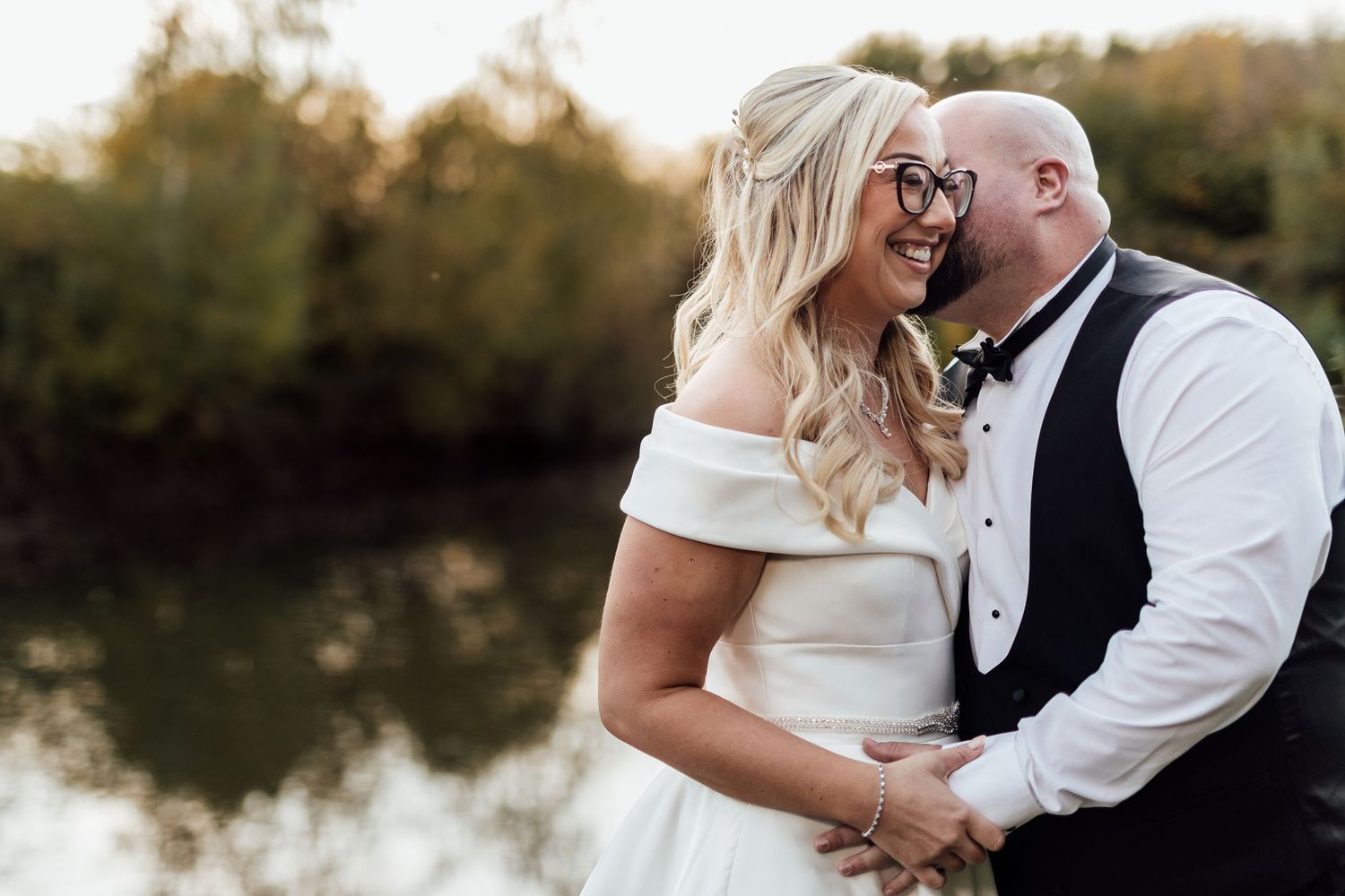 Romantic moment between newlyweds laughing together by waterside at sunset.