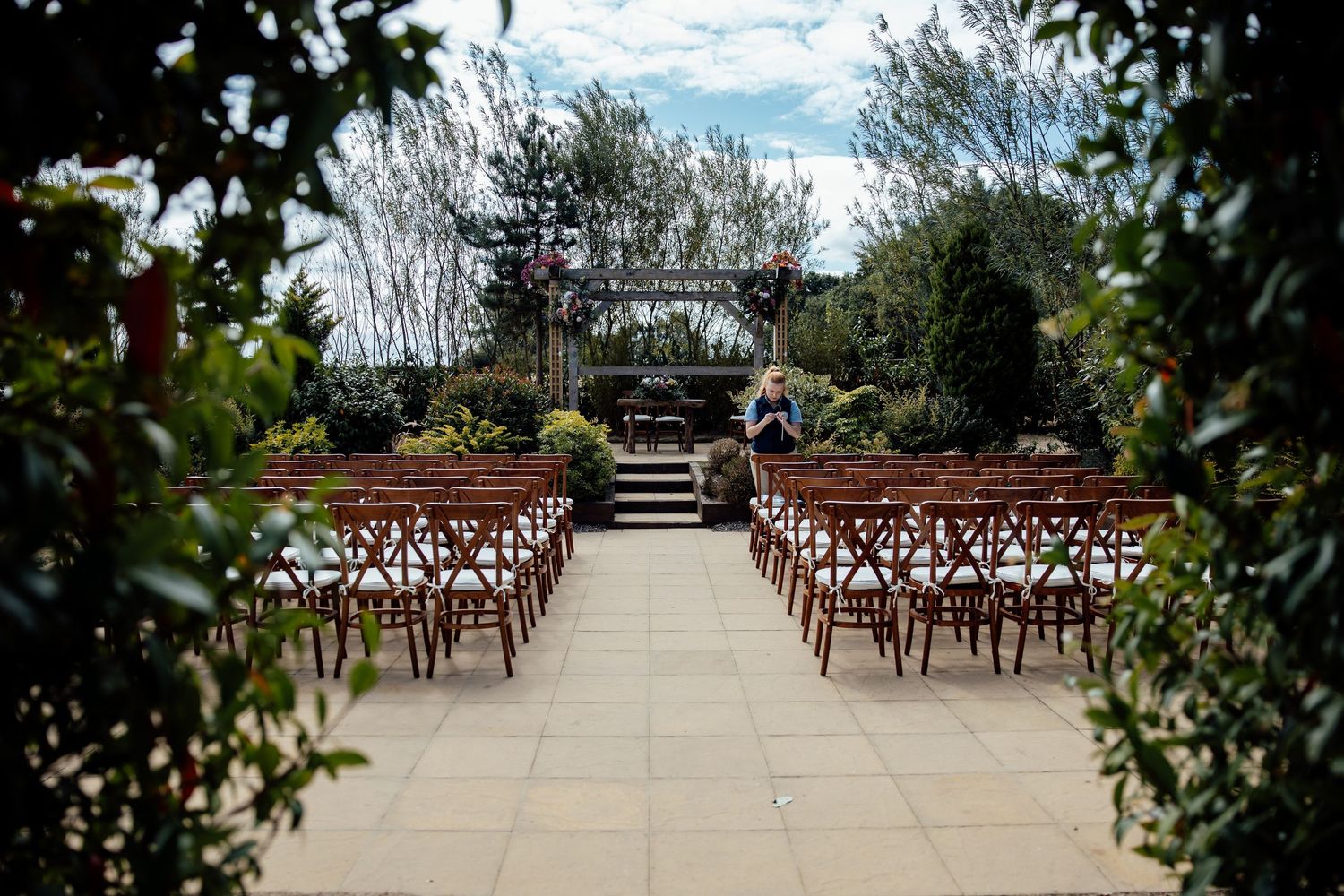 Elegant outdoor wedding ceremony setup with wooden chairs arranged in rows on a stone patio surrounded by lush greenery.