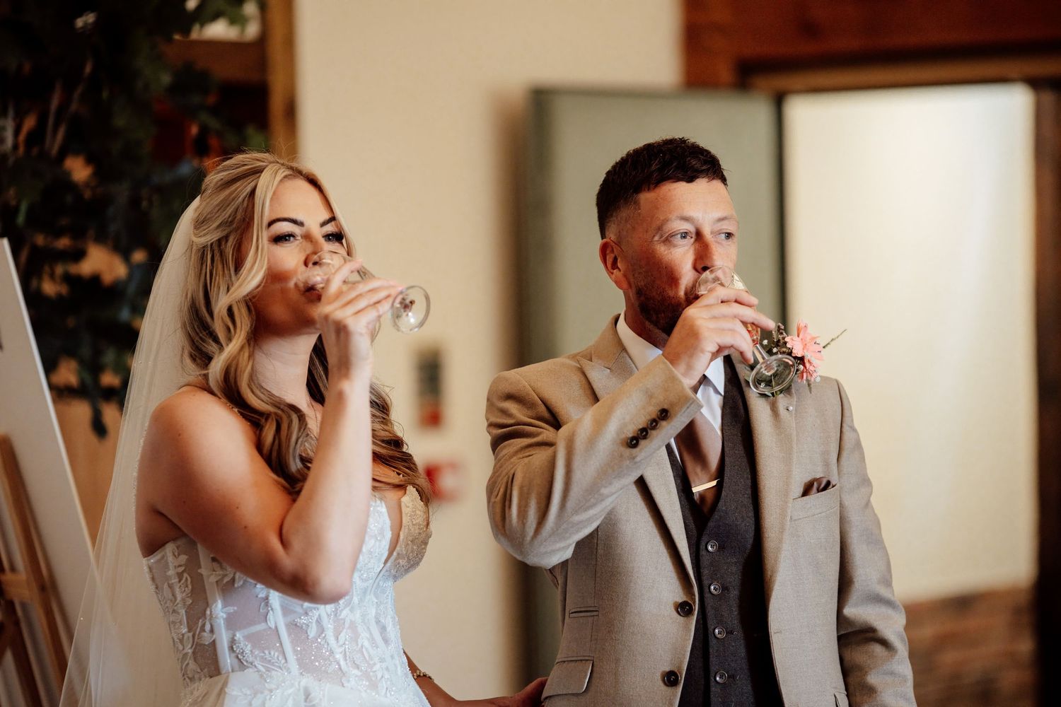 Bride in lace gown and groom in grey suit drinking during wedding reception toast ceremony.