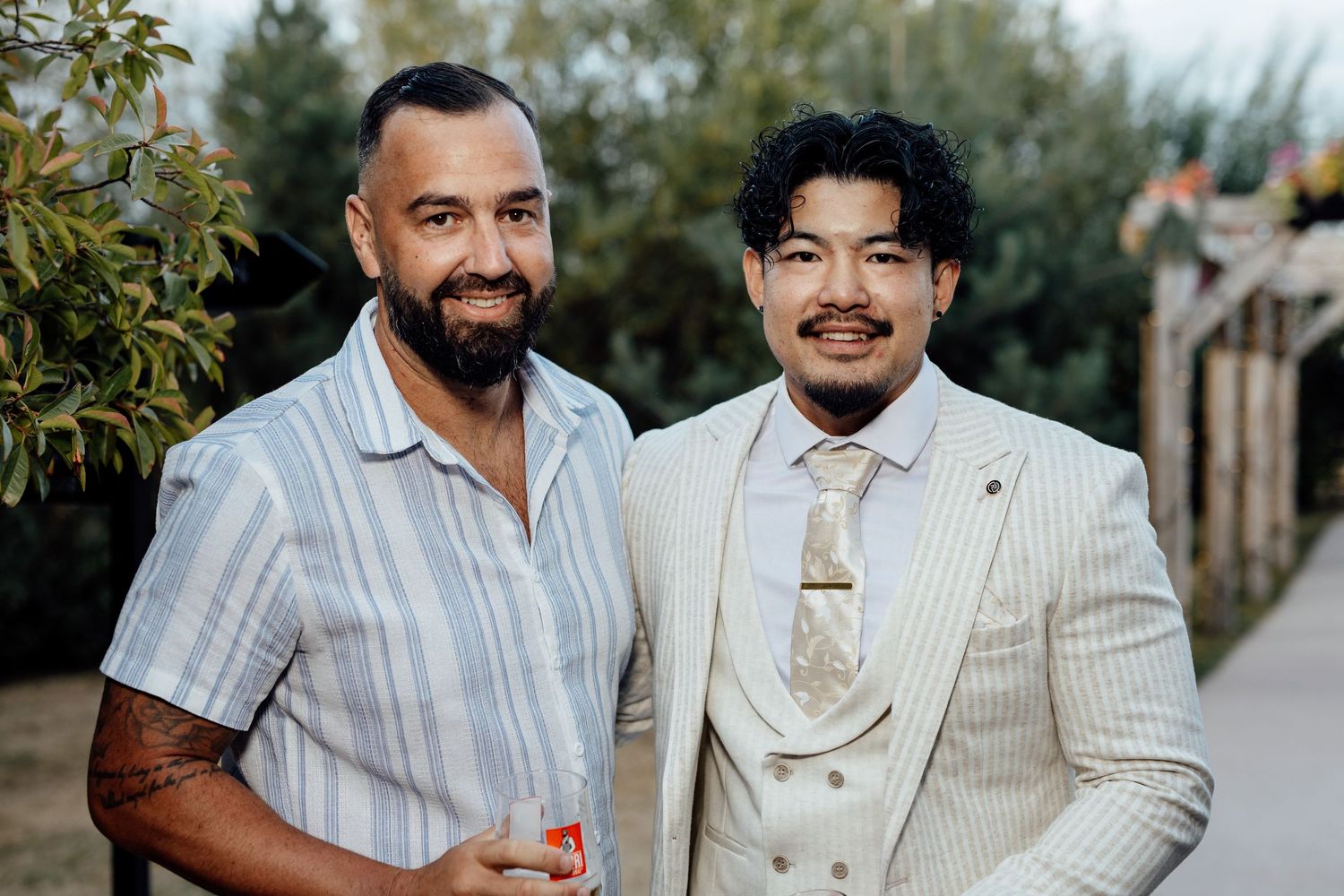 Two men smiling together outdoors at a wedding celebration in smart attire.