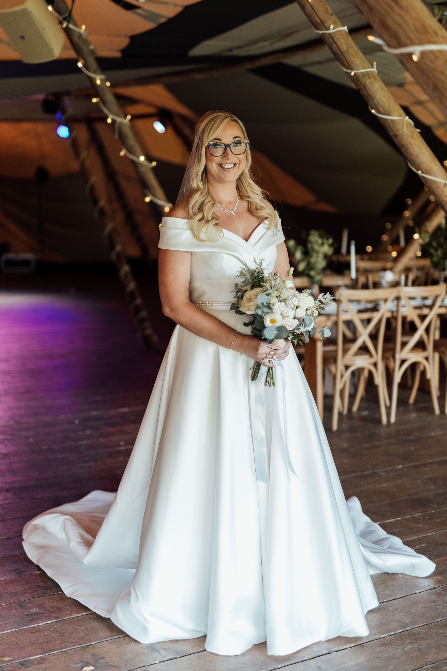 Elegant white wedding gown with off-shoulder design on wooden floor with floral bouquet.
