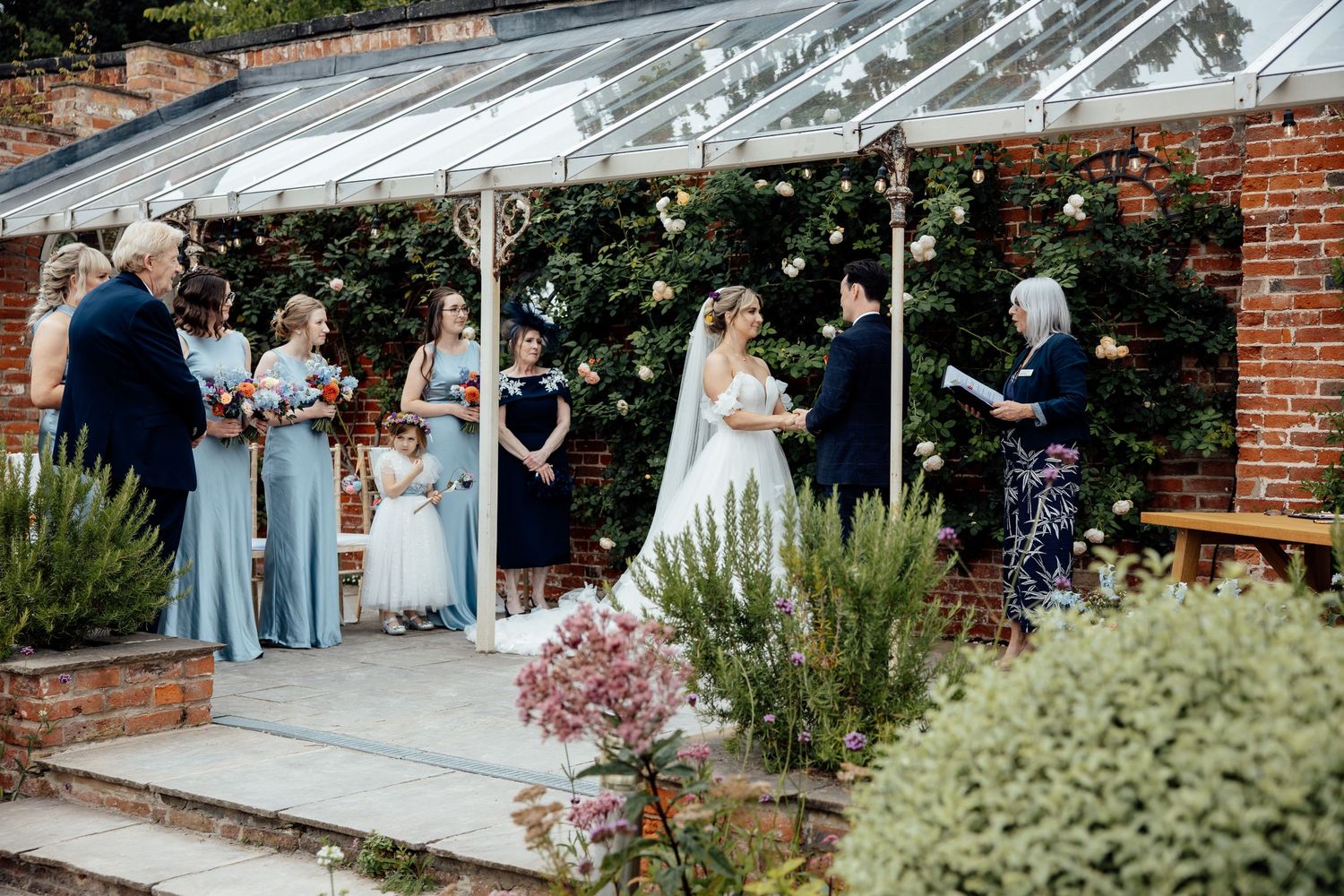 An outdoor wedding ceremony takes place under a glass-covered pergola with lush garden surroundings.