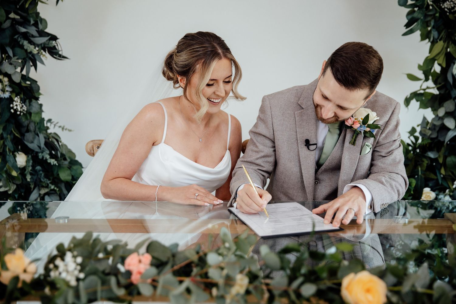 A couple signs their marriage certificate at a decorated table surrounded by floral arrangements.