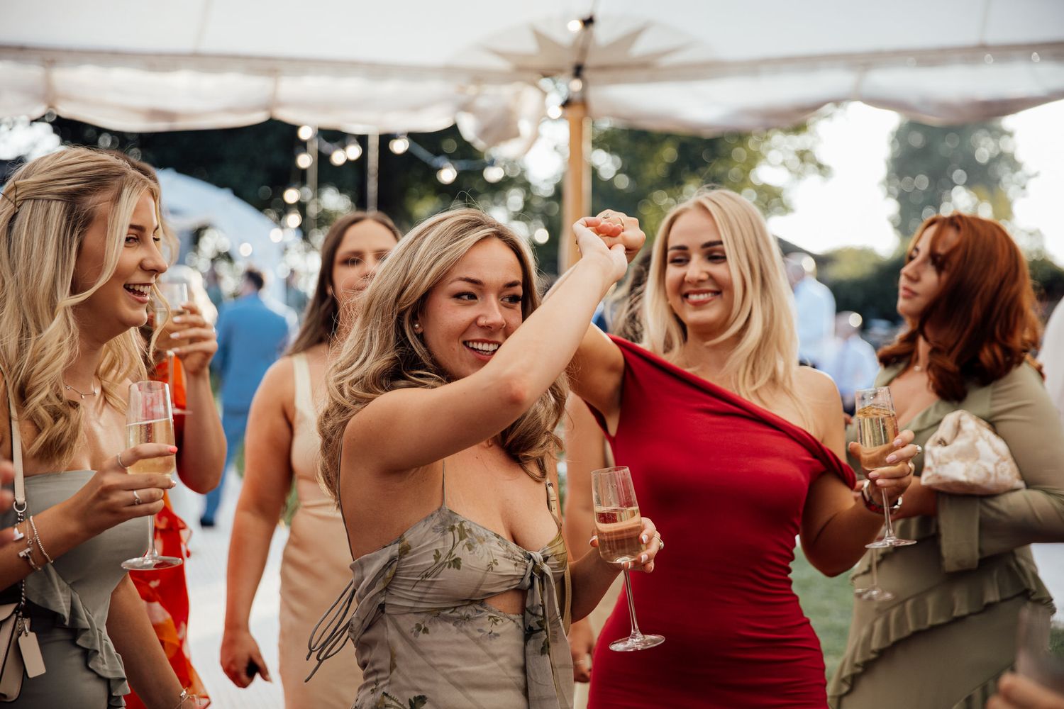 Wedding guests in elegant dresses celebrate joyfully with champagne at an outdoor reception.