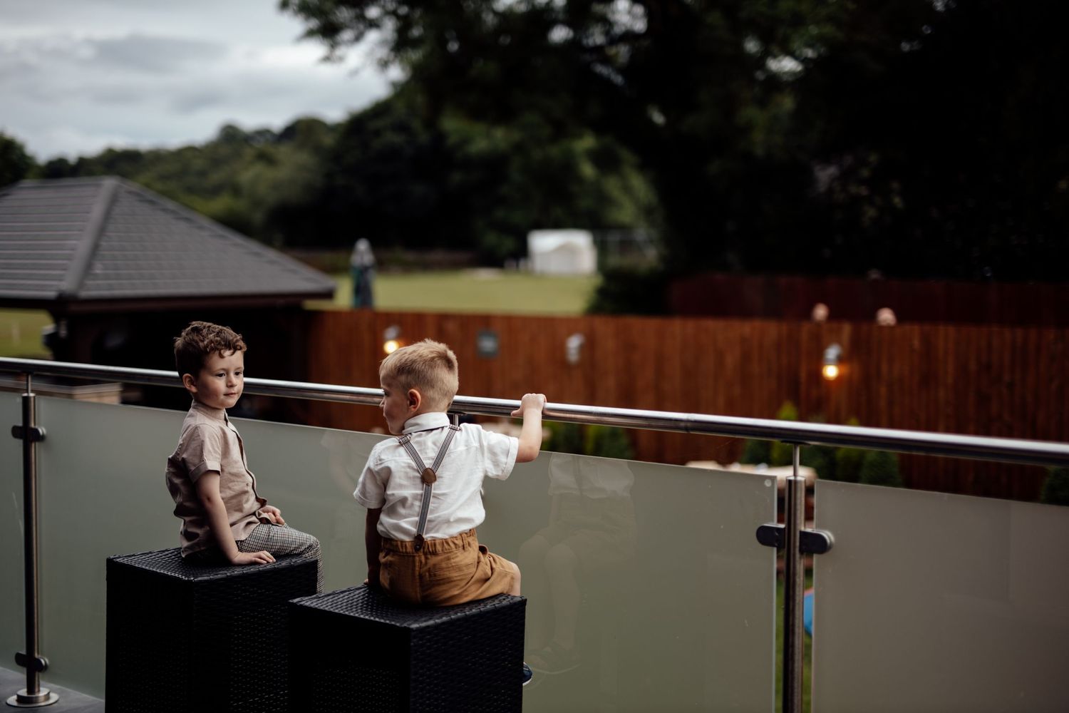 Two young children in suspenders sit on a balcony railing at an outdoor event.