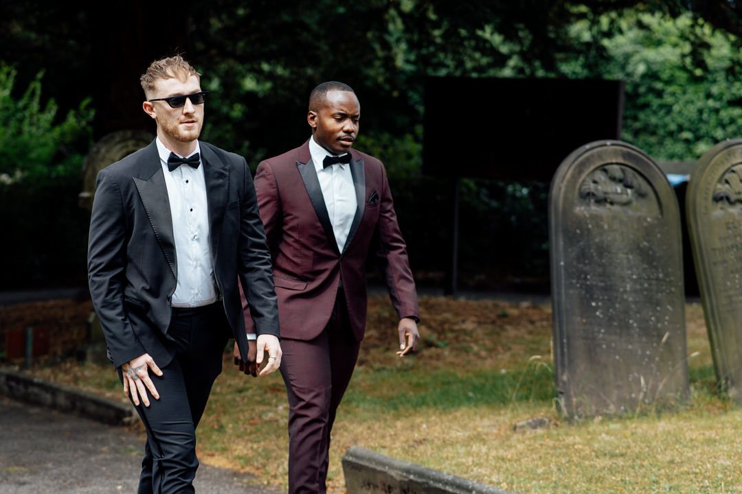 Two stylishly dressed men walking together in formal attire near cemetery grounds.