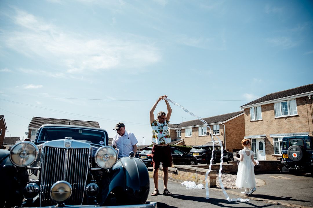 Wedding party gathered outside on a sunny day with classic cars in the background.