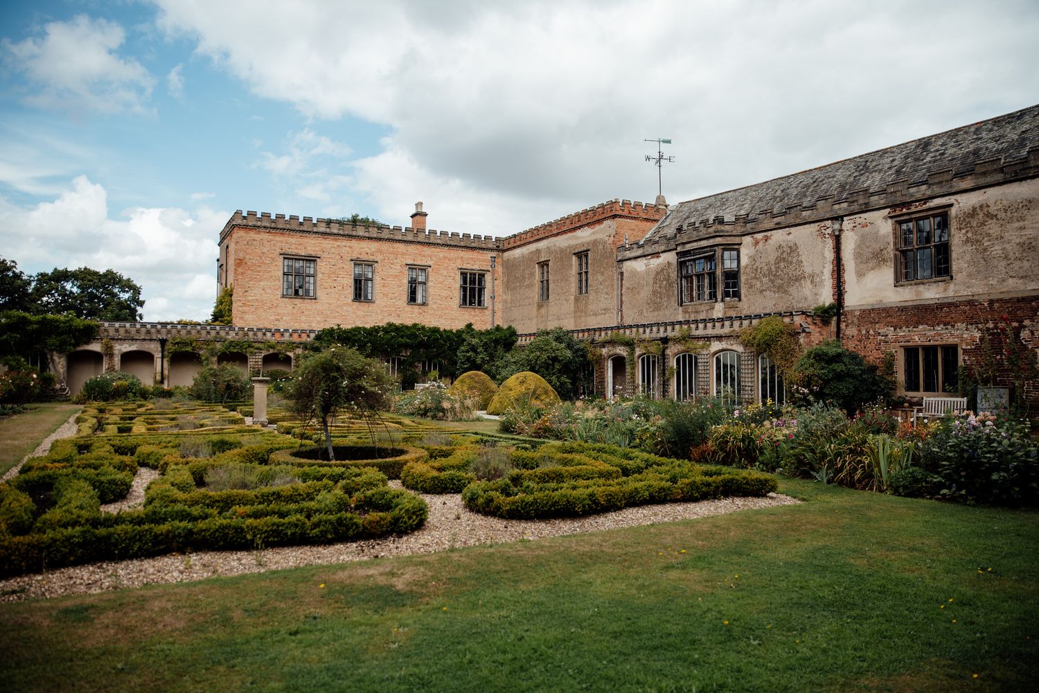 Historic stone buildings and gardens under cloudy skies create a panoramic landscape view.