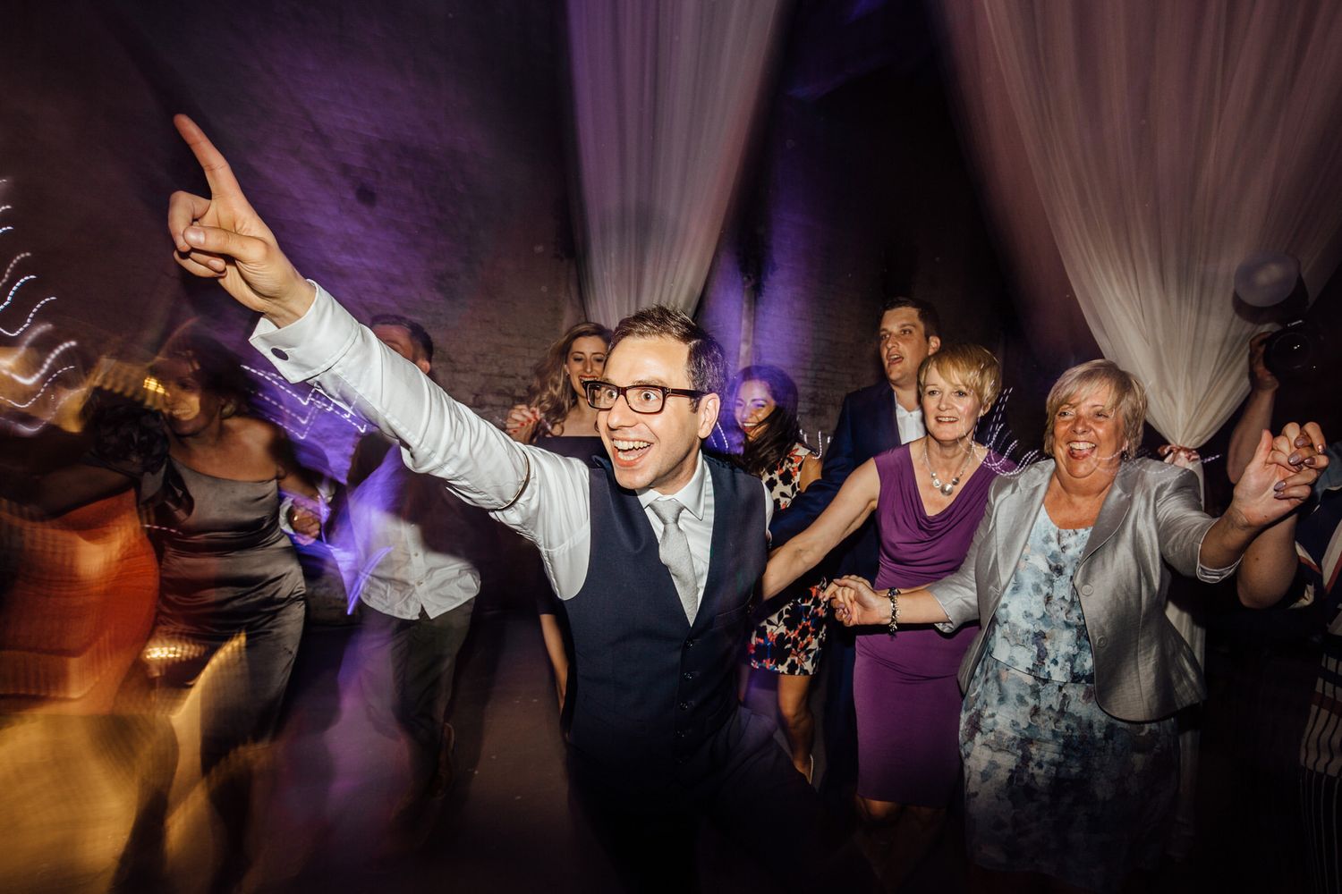 Wedding celebration on dance floor with dramatic purple lighting and white drape backdrop.