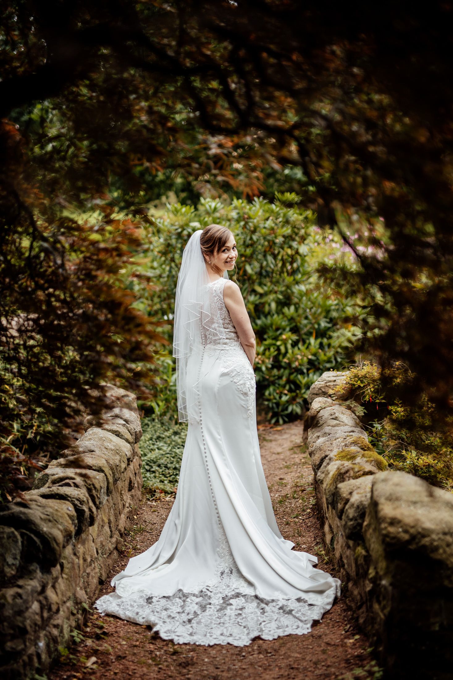 Elegant bride in lace gown and veil poses on stone path surrounded by autumn foliage.