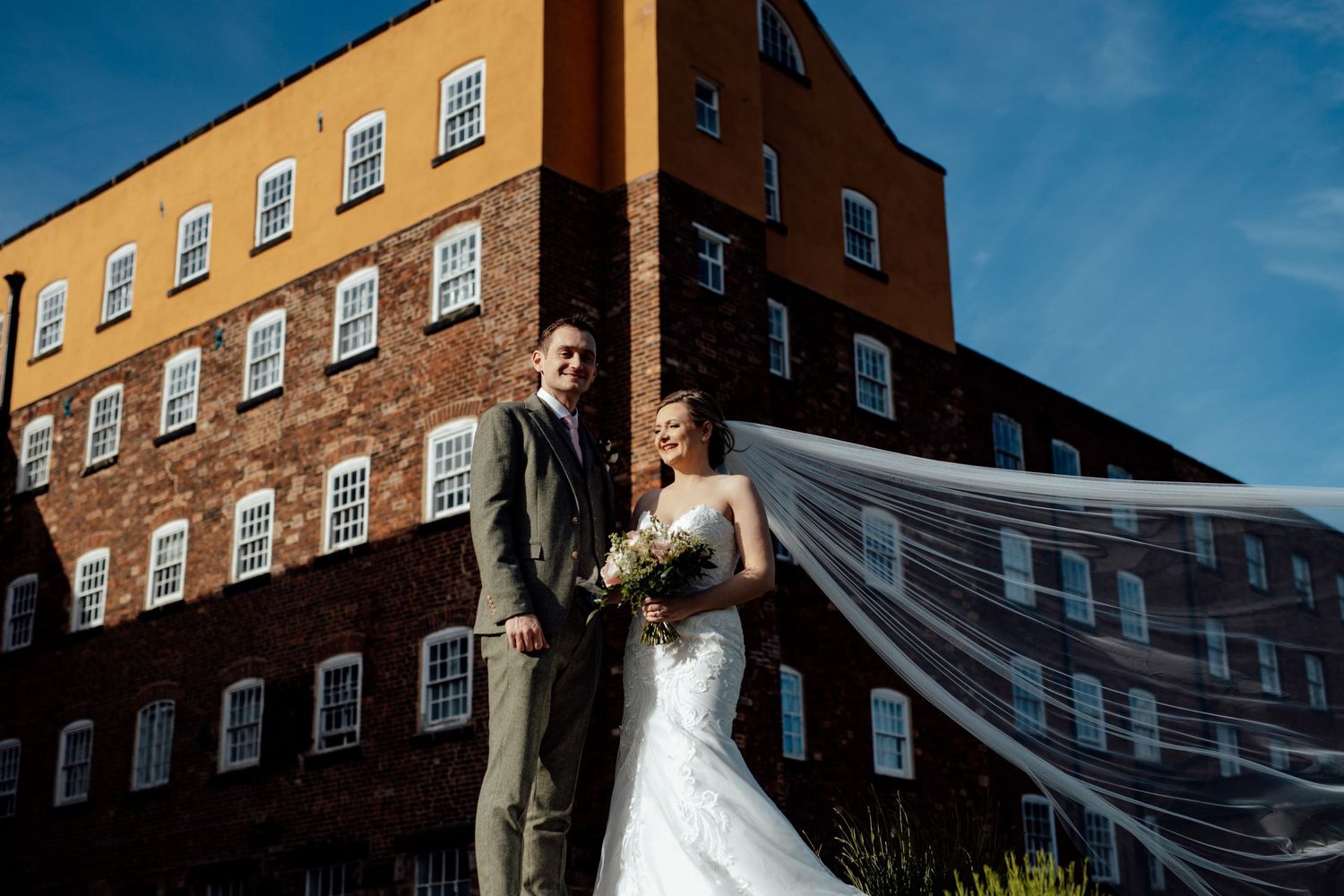A bride's veil flows dramatically in the wind against a brick building backdrop on a sunny day.