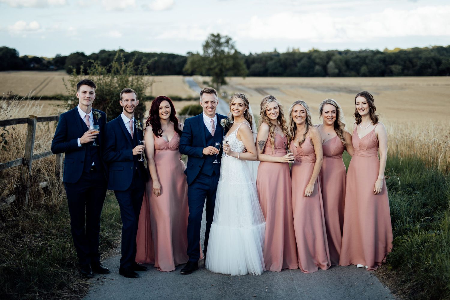 Wedding party in navy suits and dusty rose bridesmaid dresses pose together on a country path at sunset.