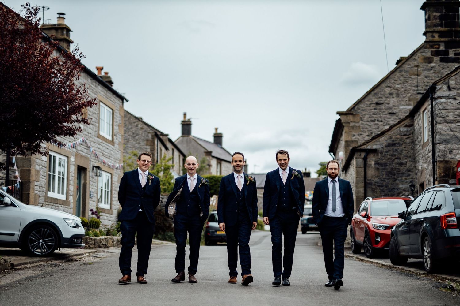 Groomsmen in dark suits walking down a village street with stone houses.