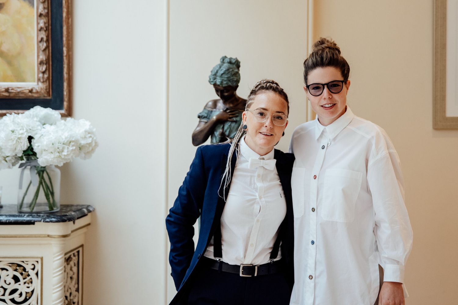 Stylish couple in formal attire posing next to an ornate side table with white flowers.