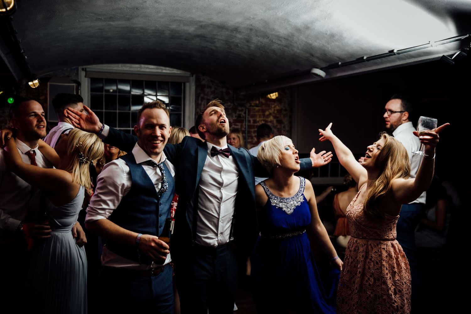 Wedding reception dance floor filled with celebrating guests in dramatic lighting.