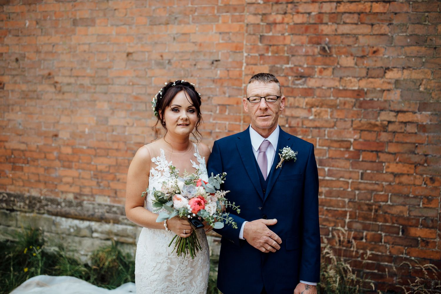 Wedding couple poses against rustic brick wall backdrop with elegant floral bouquet.