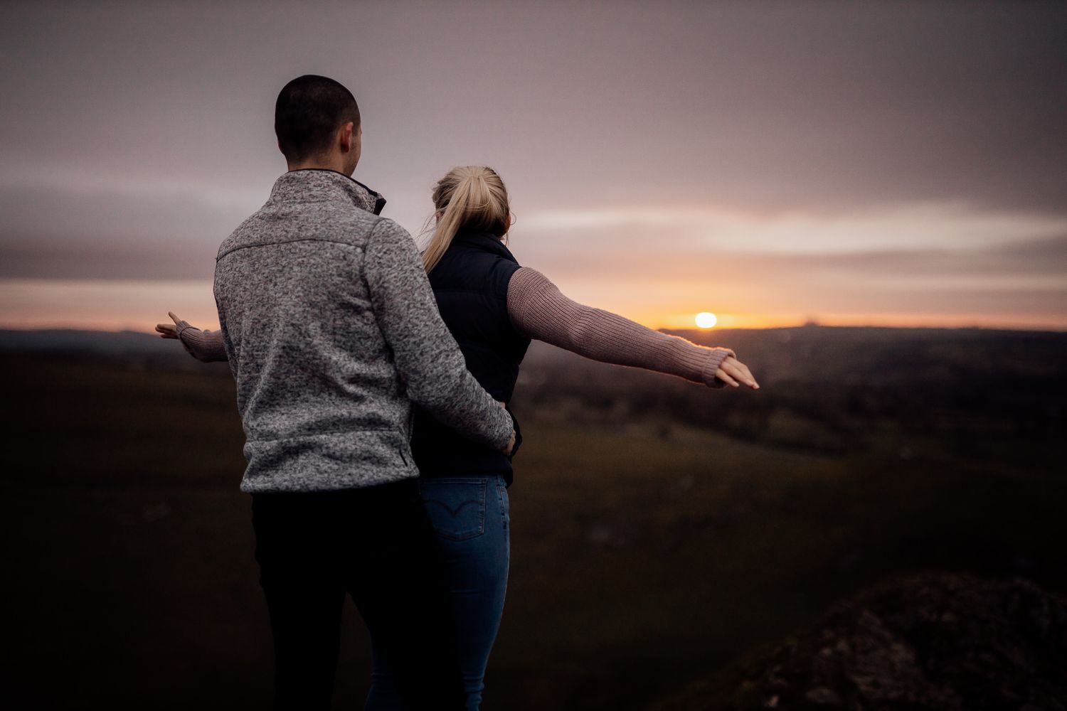 A couple embraces while watching a dramatic sunset over mountainous terrain.