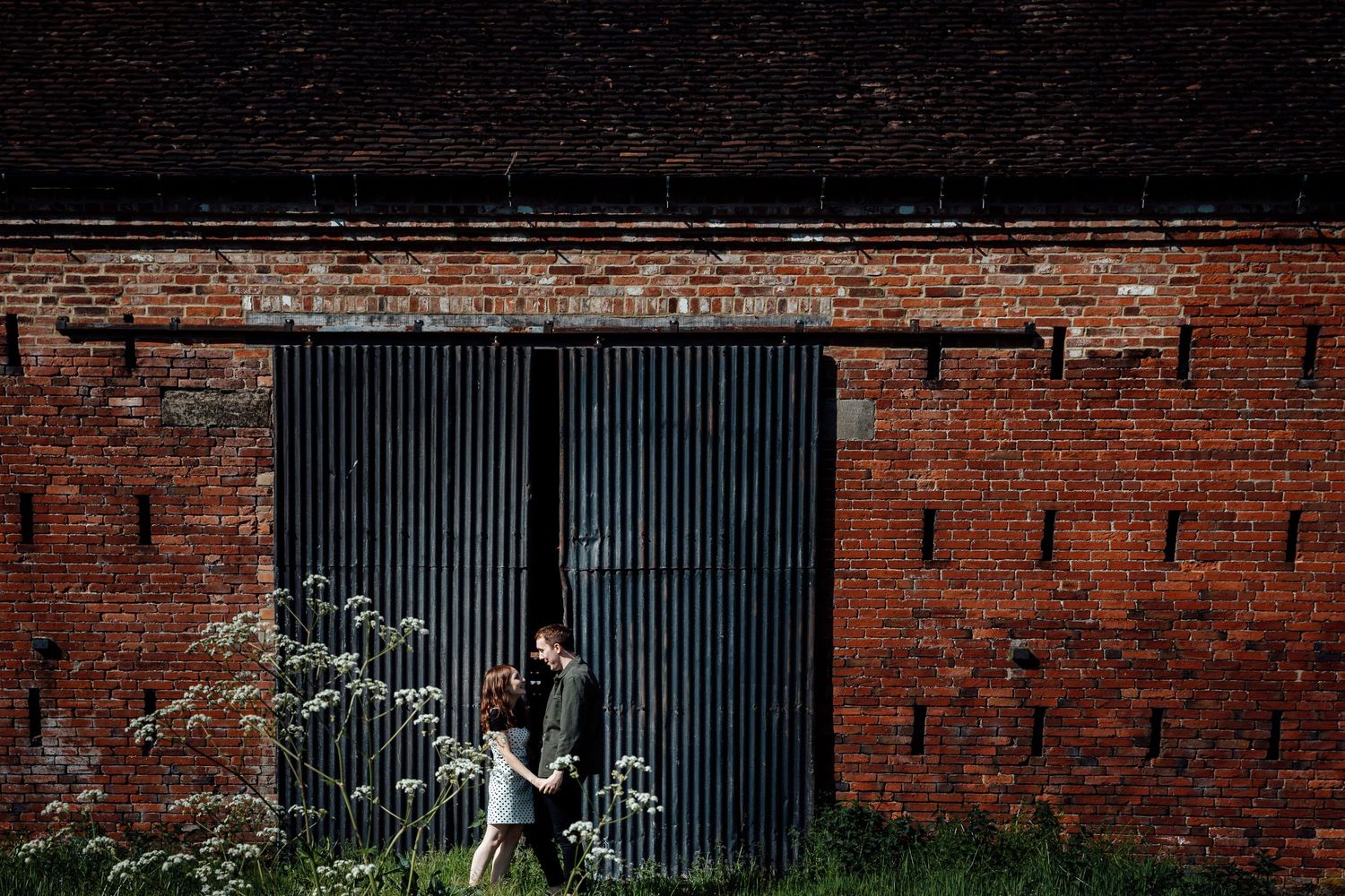 Two people standing by an industrial brick wall with metal grating.