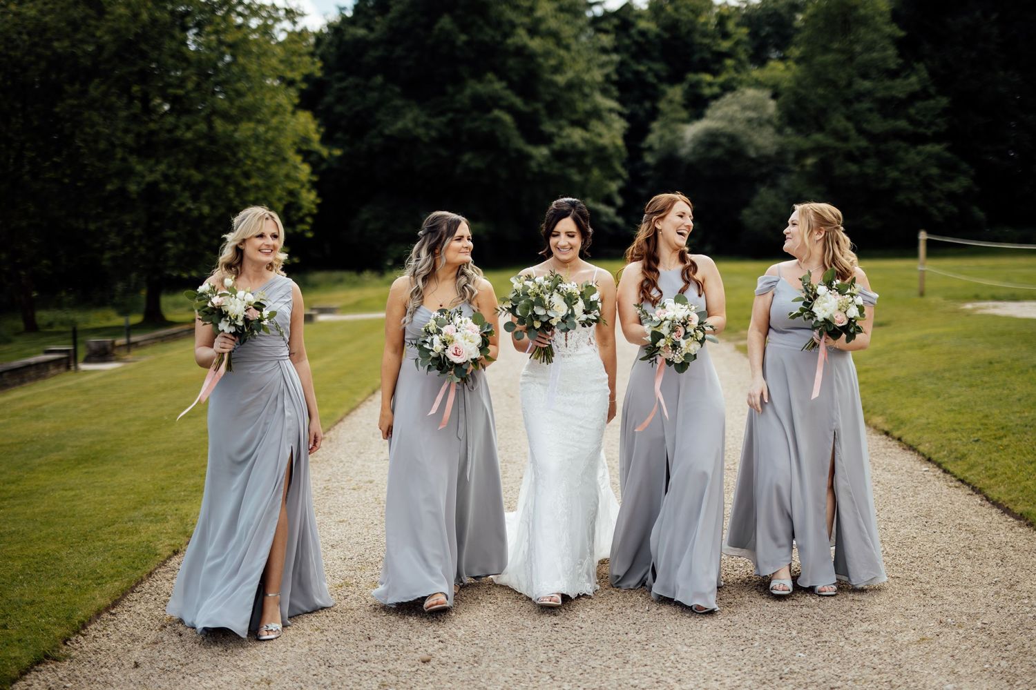 Bridesmaids in flowing gray dresses walking together on a garden path holding bouquets.