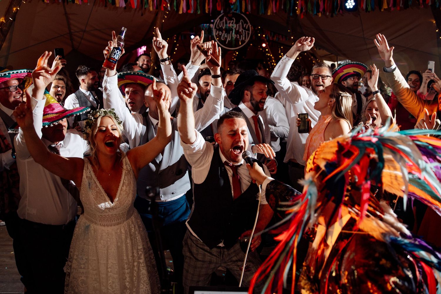 Crowd of wedding guests dancing with raised hands under colorful bunting at a lively reception celebration.