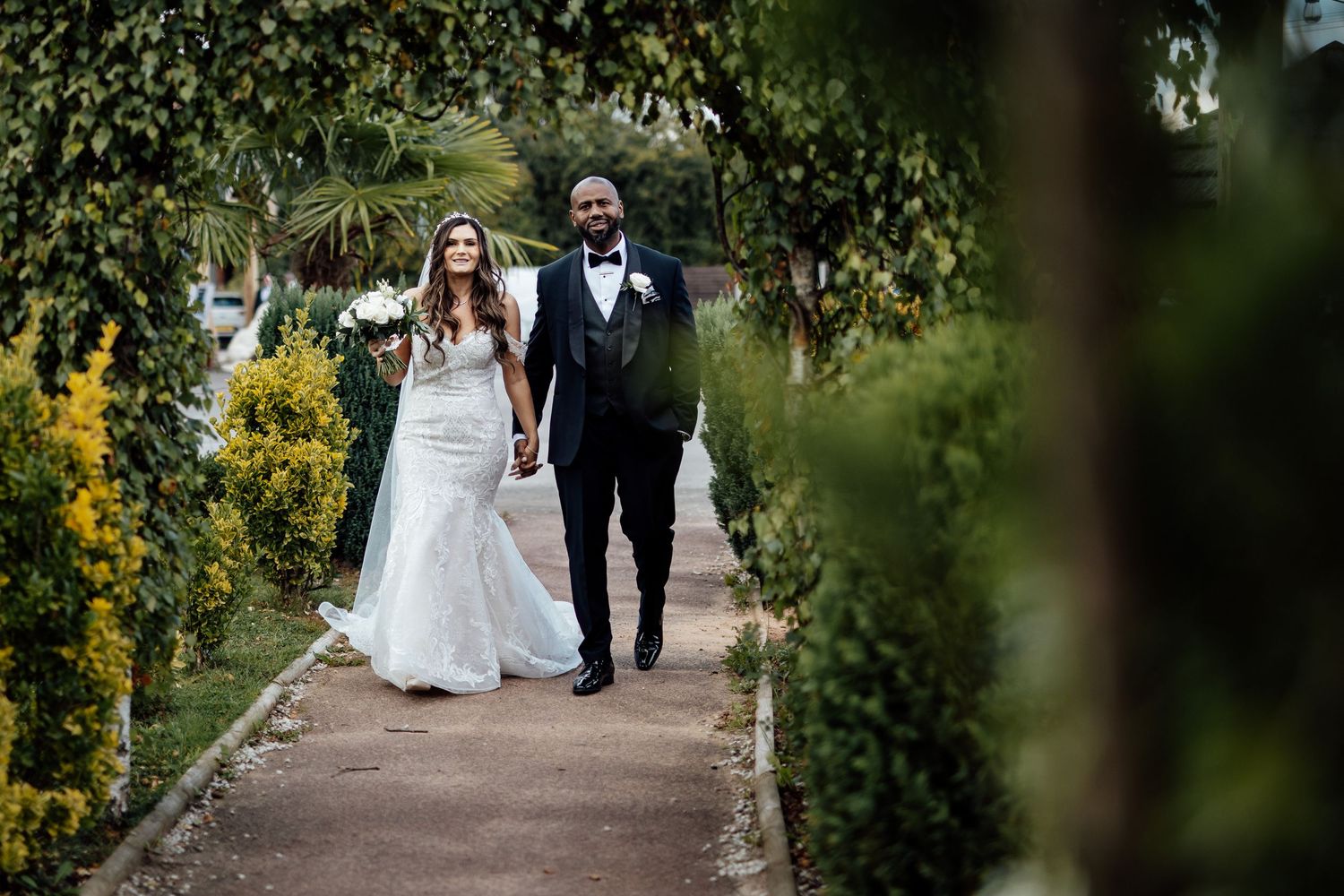 A couple walks along a garden path surrounded by lush greenery and yellow flowers on their wedding day.