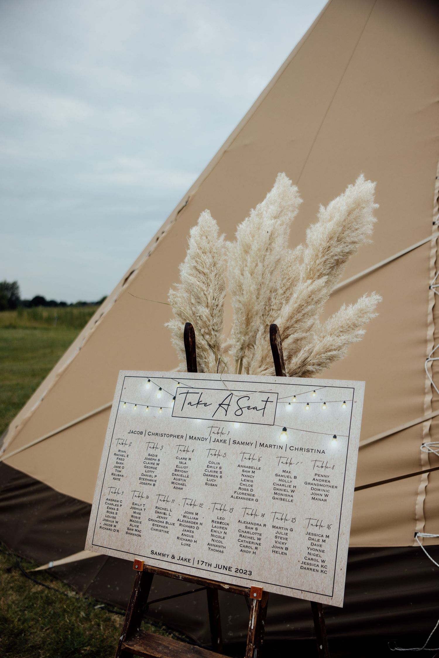 Wedding seating chart displayed outside a beige tipi tent with pampas grass decorations.