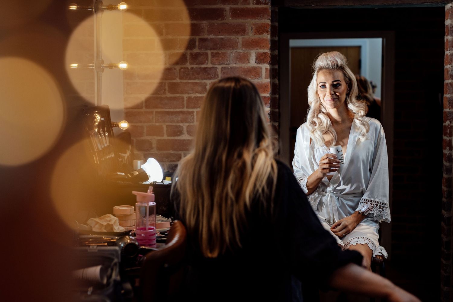Intimate getting ready moments with soft bokeh lighting and brick wall background.