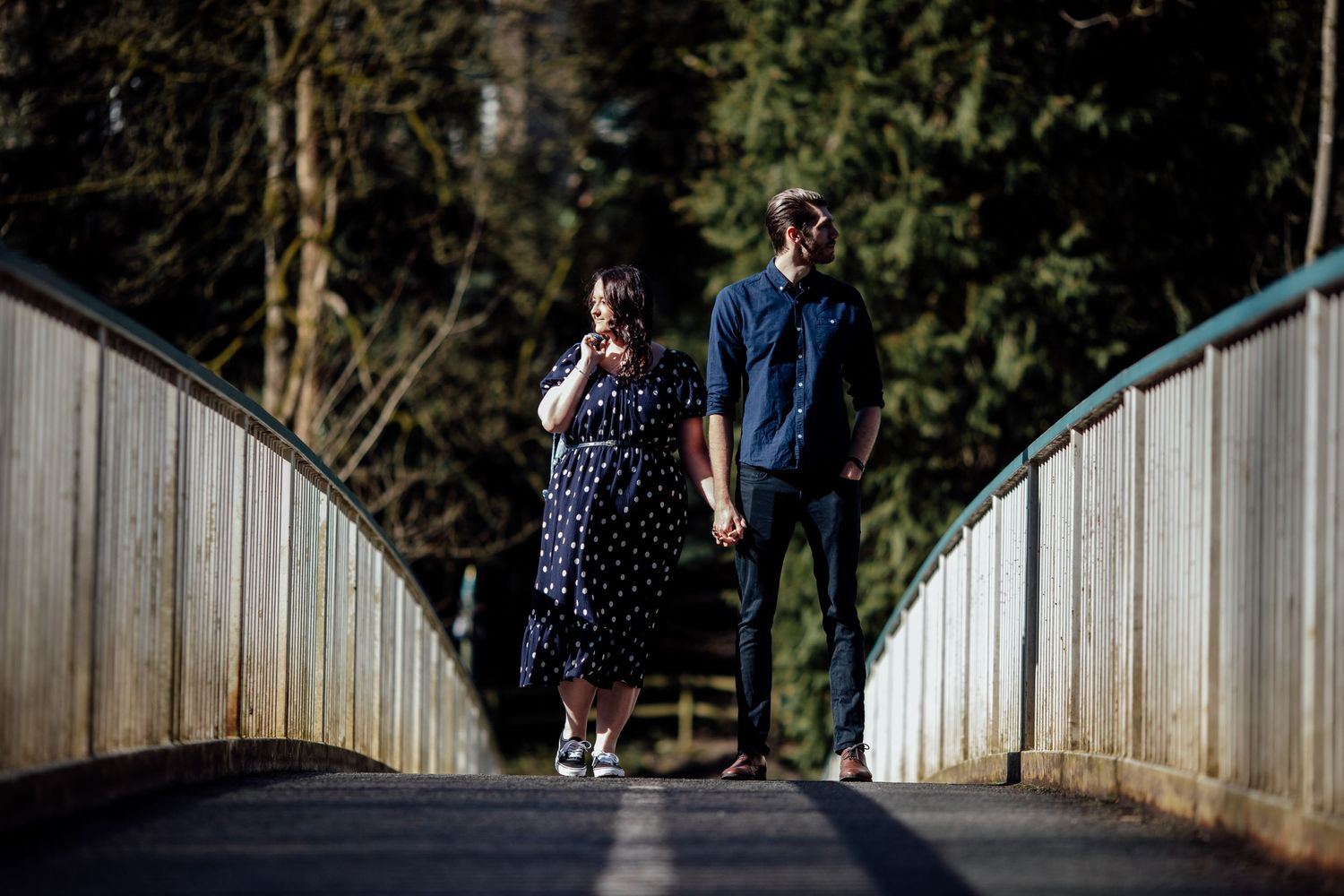 A couple stands on a wooden bridge surrounded by forest in dramatic lighting.