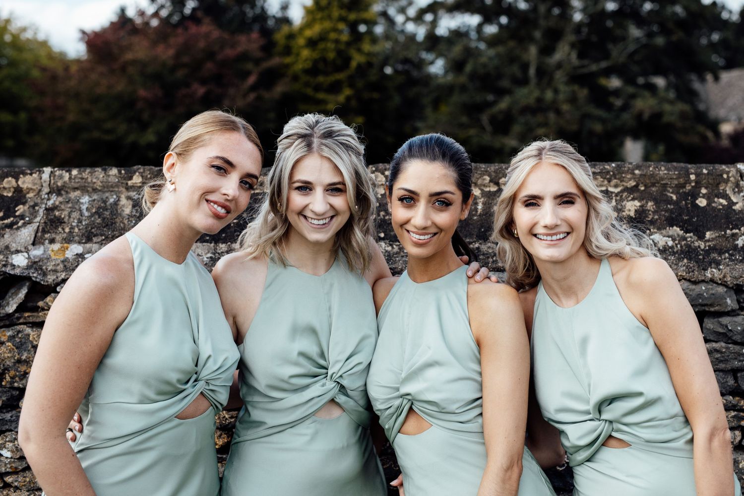 Bridesmaids wearing sage green dresses pose together outdoors for wedding photos.