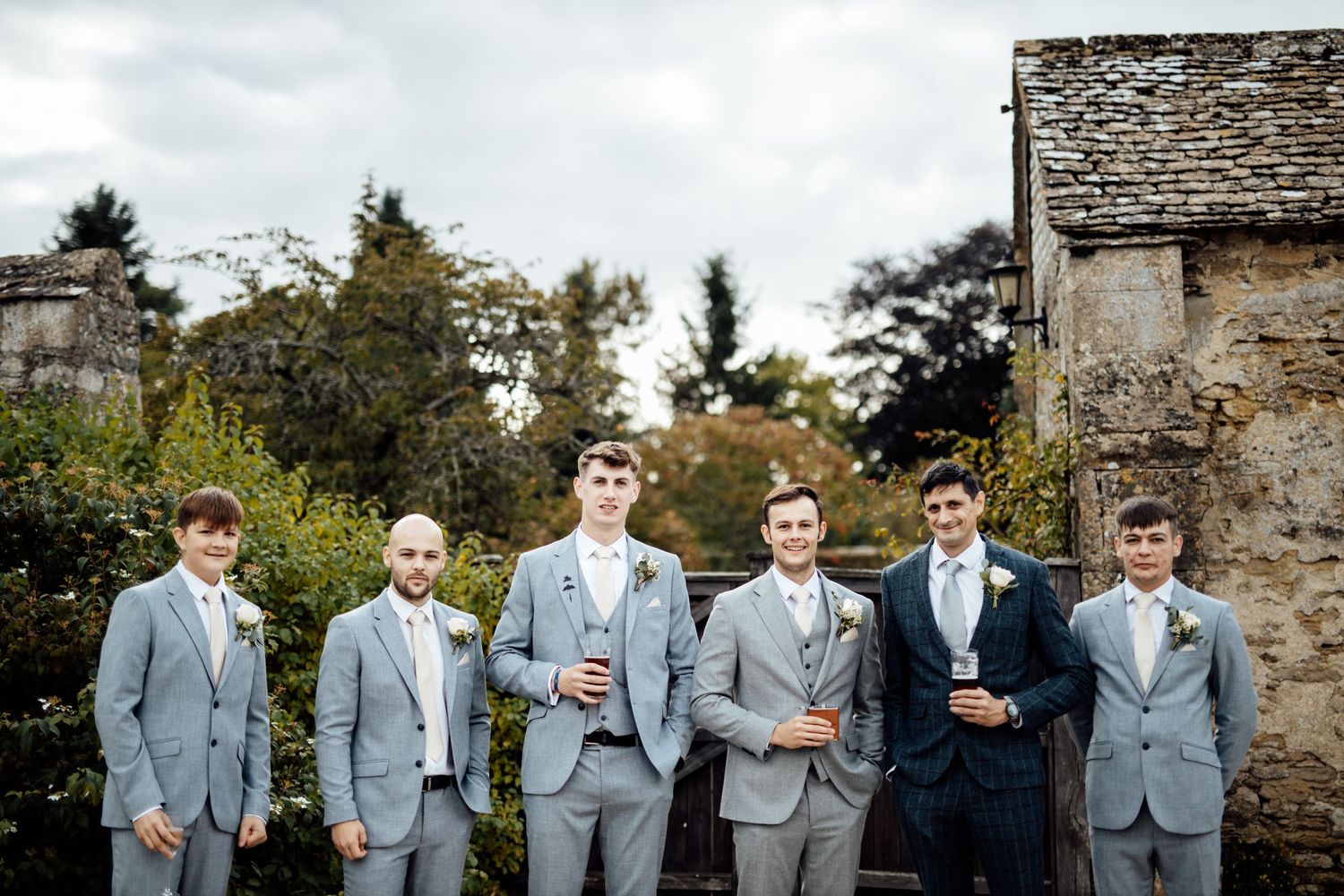 Groomsmen in light grey suits gather for a photo against a stone wall backdrop.