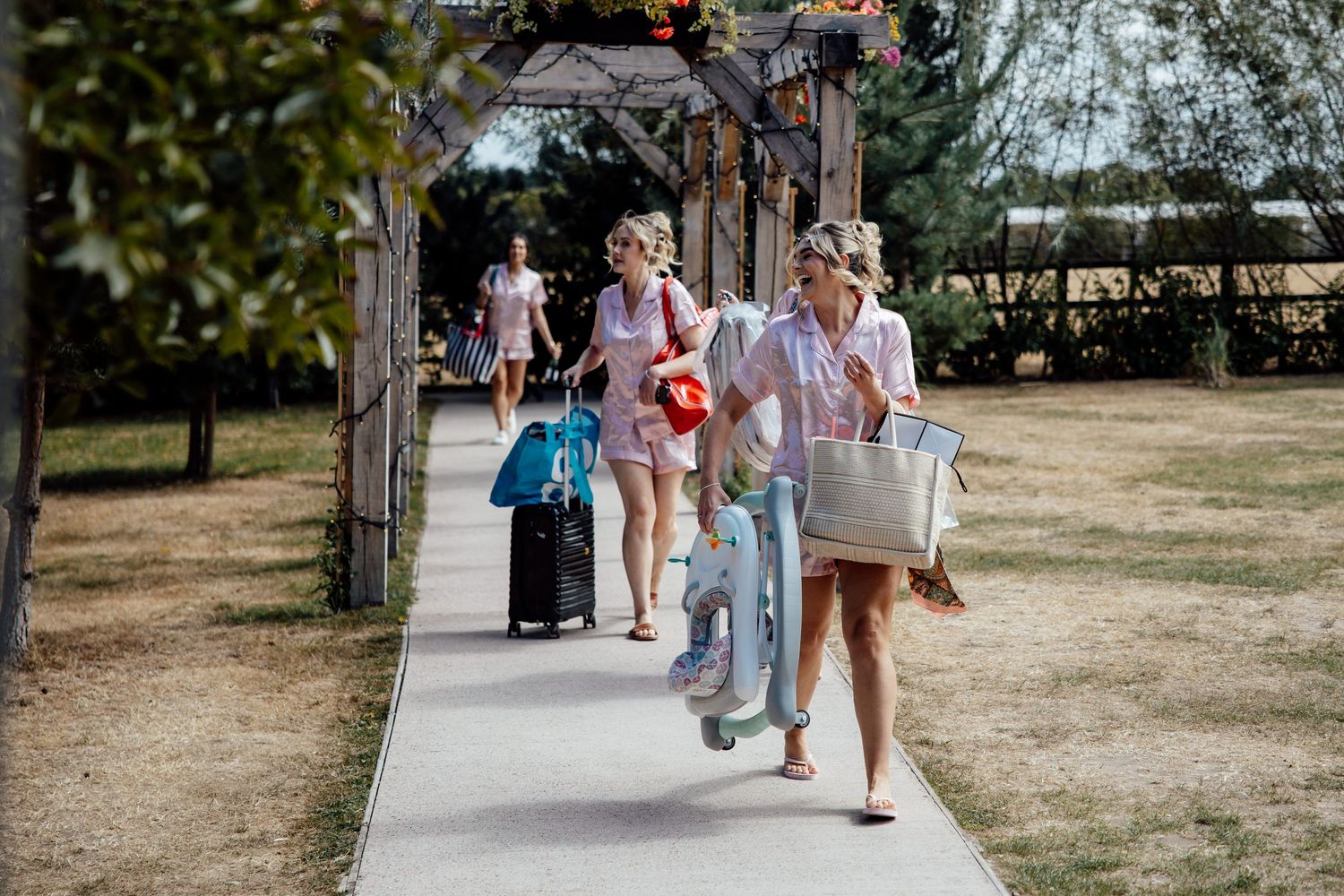 Women walking along a garden pathway carrying luggage and bags on a sunny day.
