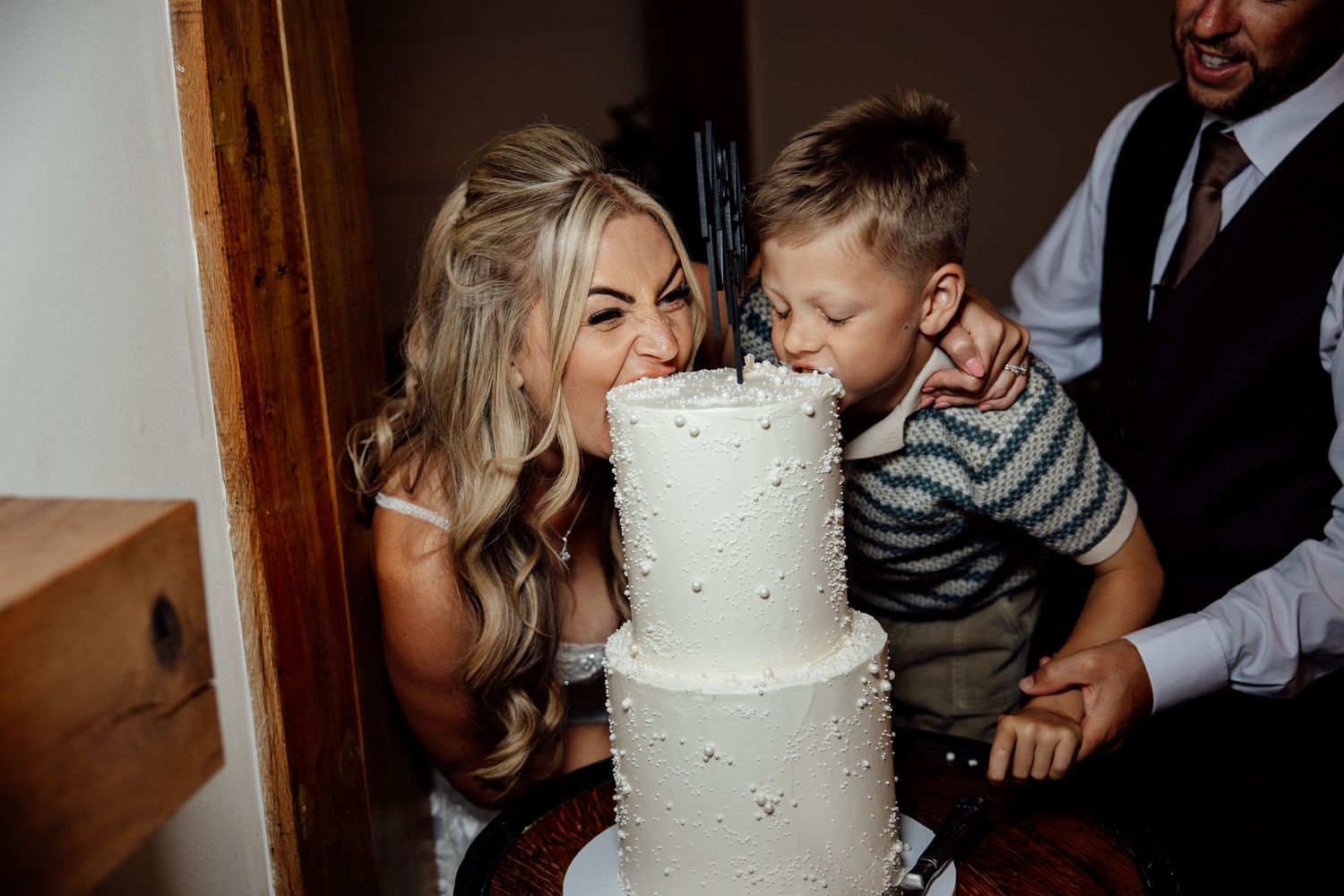 Bride and young boy playfully bite into a white three-tier wedding cake together at reception.