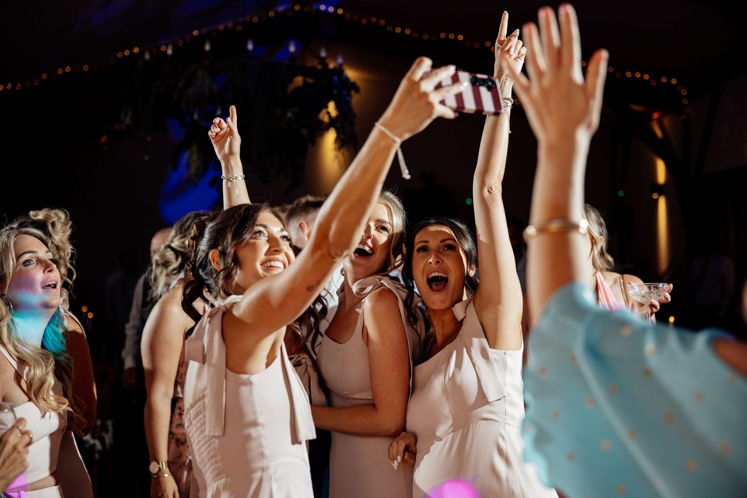 Bridesmaids in white dresses dancing and cheering with hands raised at wedding reception.