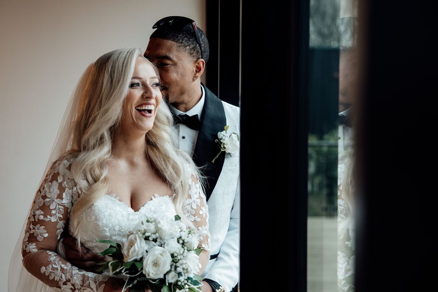 Laughing bride in lace gown holding white bouquet with groom whispering in her ear by a window.