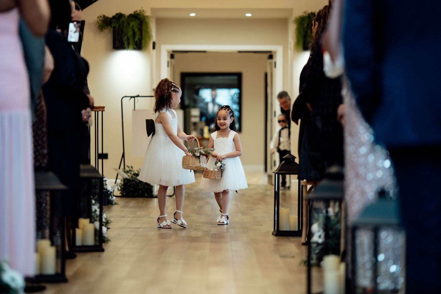 Two flower girls in white dresses walk down the wedding aisle carrying a basket of flowers.