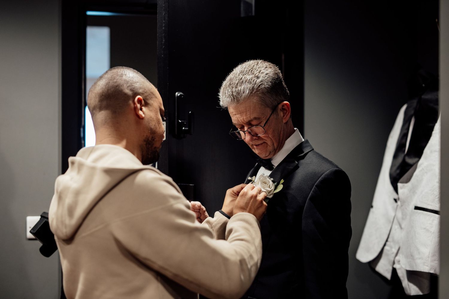 Groom having boutonniere pinned to his black suit lapel by a man in a beige hoodie in a dark room.