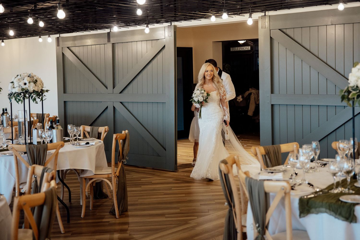 Bride in lace wedding gown enters elegant barn reception venue through large blue doors.