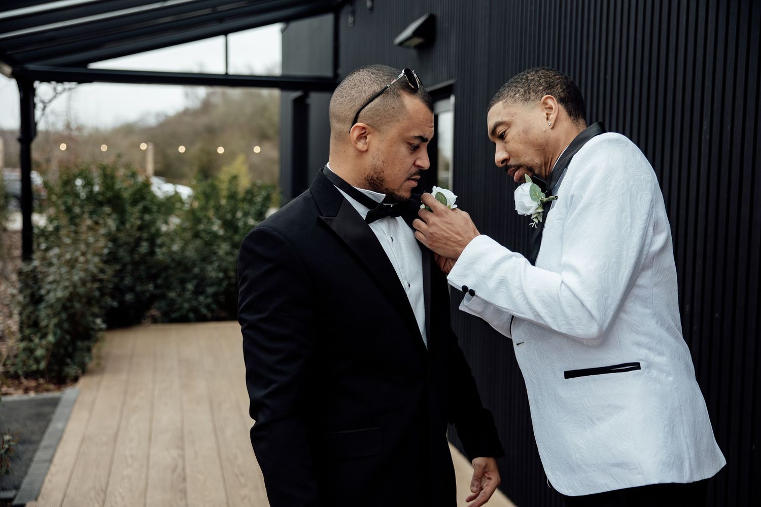 Two grooms in black and white tuxedos adjusting boutonnieres outdoors before wedding ceremony.