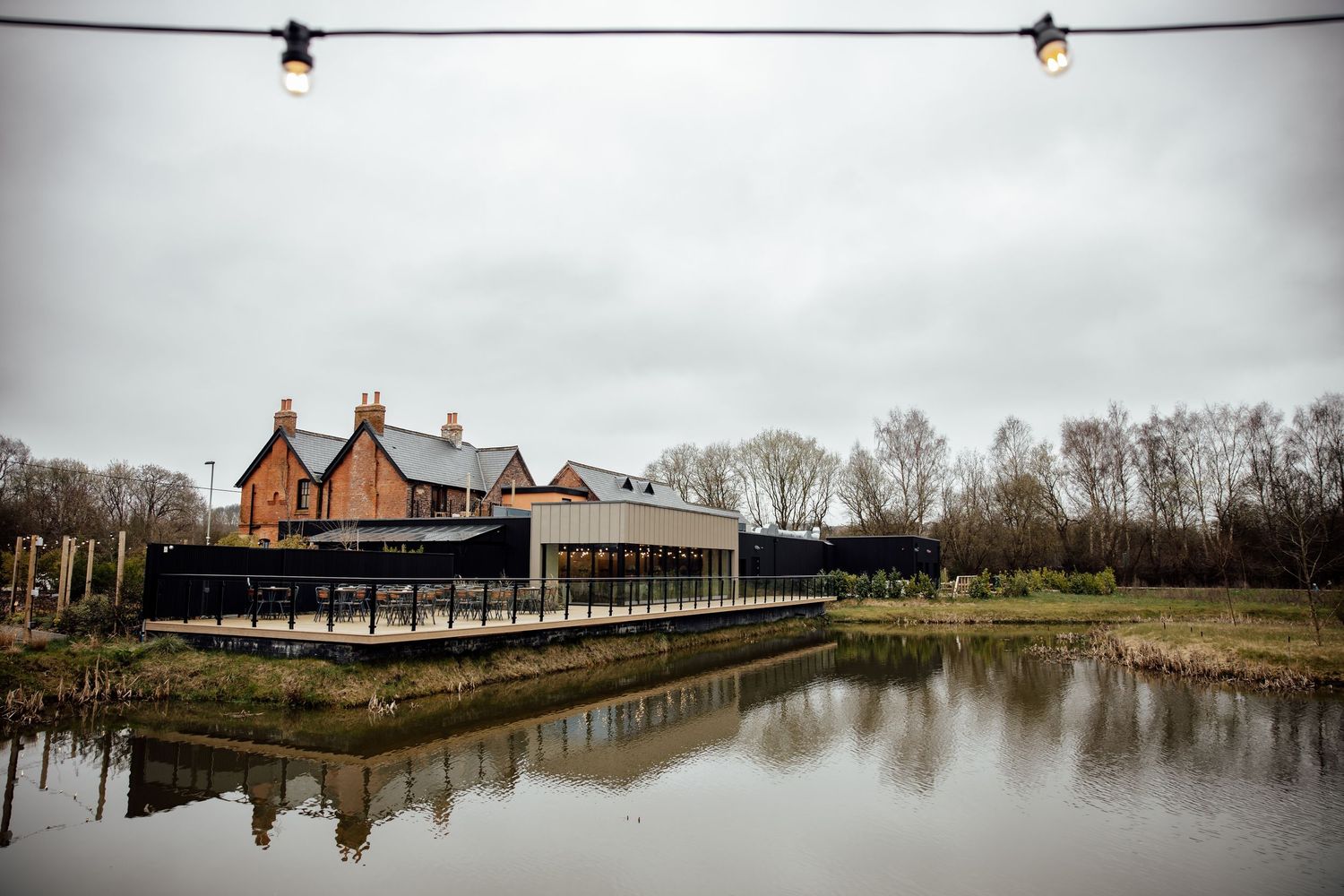 Rustic wedding venue with brick building beside a tranquil pond, surrounded by bare winter trees under an overcast sky.