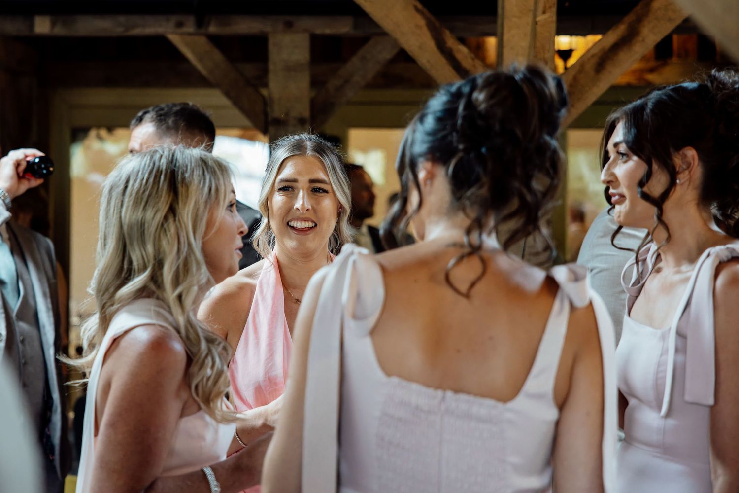 Bridesmaids in pink dresses laughing and chatting with bride inside rustic wooden barn wedding venue.