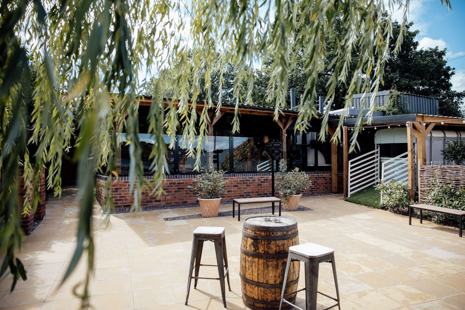 Rustic outdoor wedding venue patio with barrel tables, stools, and weeping willow tree.