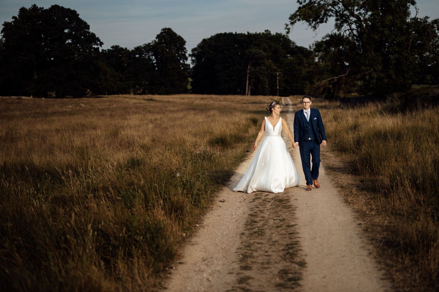 A couple walks hand in hand down a dirt path through a golden field at sunset.