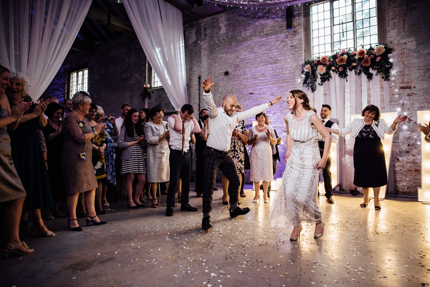 Wedding party dancing and celebrating with confetti in a dramatically lit brick venue.